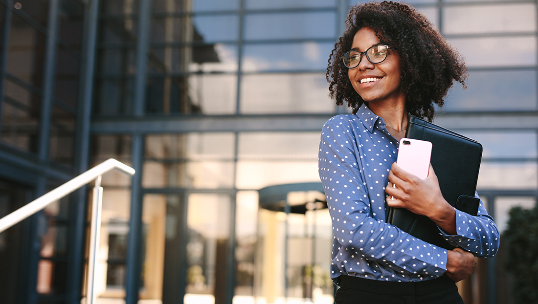 Graduate development: a woman with mobile phone and file standing outside a building.