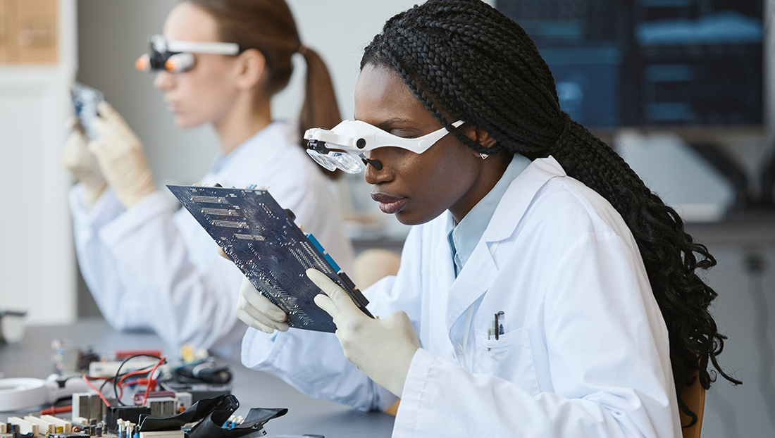 STEM graduates: Black woman wearing magnifying glasses and inspecting electronic parts in quality control lab